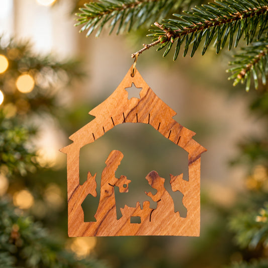 A wooden nativity ornament shaped like a stable hangs from a pine branch, with a blurred Christmas tree and warm lights in the background.