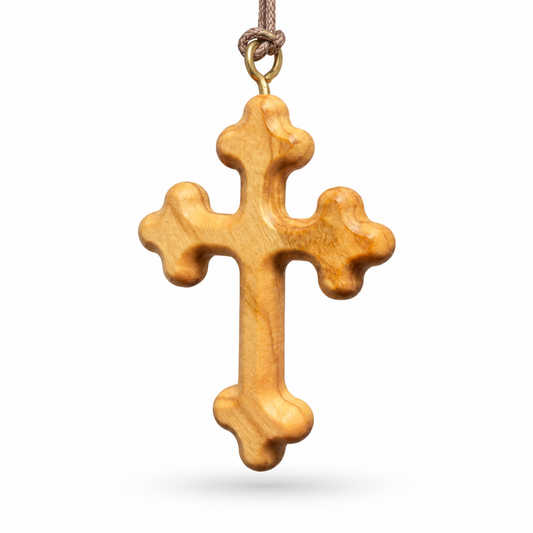 A carved olive wood cross pendant with a natural warm tan-brown finish, hanging from a brown cord with a brass-colored loop, shown against a plain white background.