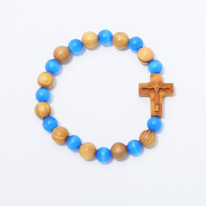A beaded bracelet featuring alternating light brown wooden beads and bright blue acrylic beads, with a small carved wooden cross on the right side, displayed on a clean white background.