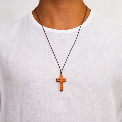 A close-up of a person wearing a brown wooden cross pendant necklace on a dark cord, paired with a white linen t-shirt against a light background.