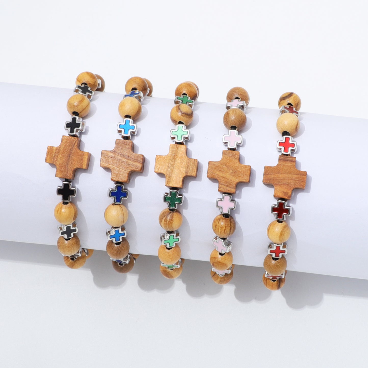 Five wooden beaded bracelets with cross charms are displayed on a white cylindrical stand against a white background. The crosses feature different colored enamel details in black, blue, green, pink, red, and white, paired with light brown and beige wooden beads.