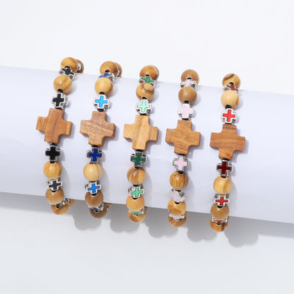 Five wooden beaded bracelets with cross charms are displayed on a white cylindrical stand against a white background. The crosses feature different colored enamel details in black, blue, green, pink, red, and white, paired with light brown and beige wooden beads.
