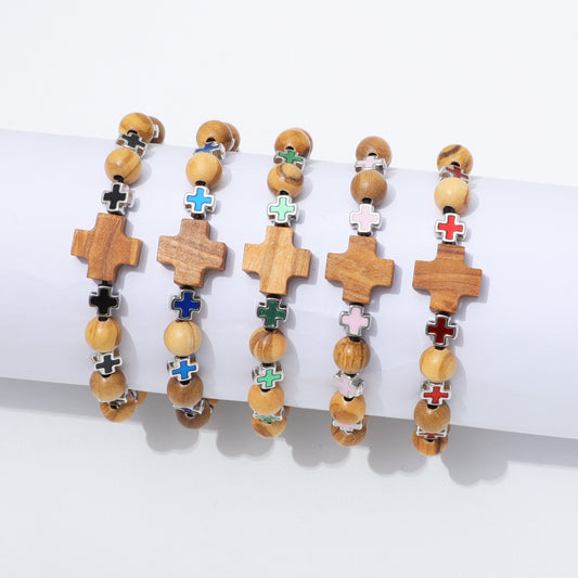 Five wooden beaded bracelets with cross charms are displayed on a white cylindrical stand against a white background. The crosses feature different colored enamel details in black, blue, green, pink, red, and white, paired with light brown and beige wooden beads.