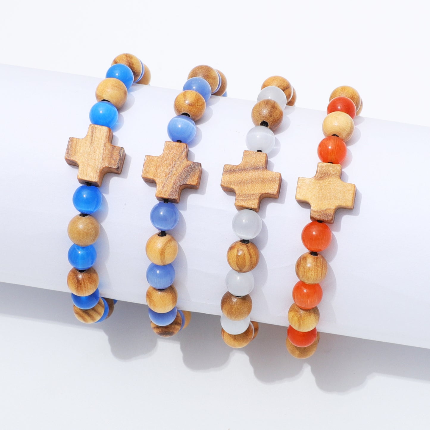 Four beaded bracelets with wooden cross charms displayed on a white cylindrical stand. The bracelets feature natural wood, blue beads, white beads, and orange carnelian beads.
