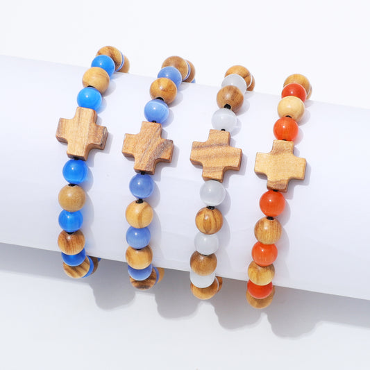 Four beaded bracelets with wooden cross charms displayed on a white cylindrical stand. The bracelets feature natural wood, blue beads, white beads, and orange carnelian beads.