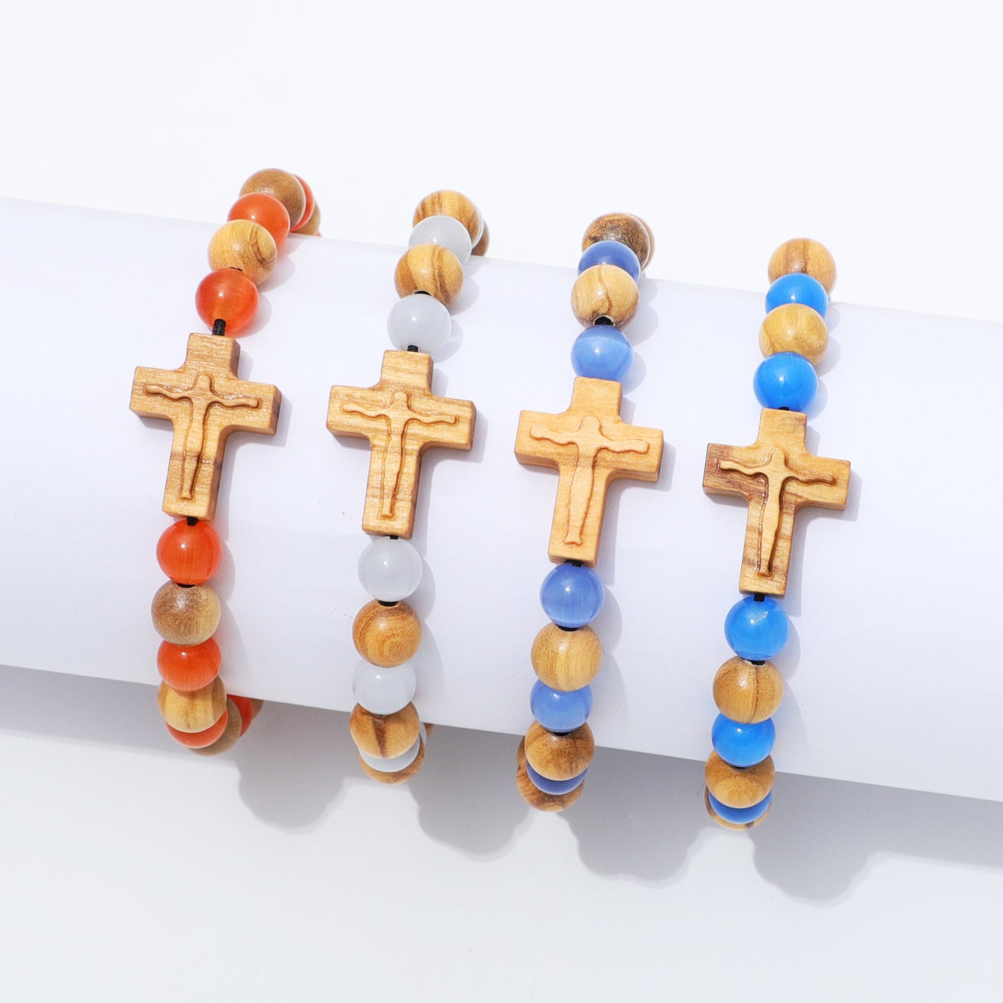 Four beaded bracelets with wooden cross charms displayed on a white cylindrical stand. The bracelets feature natural wood beads, orange round beads, translucent white beads, and bright blue beads.