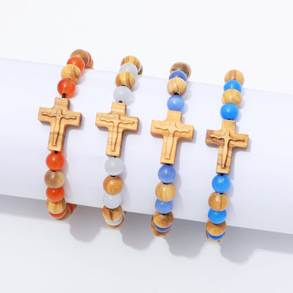 Four beaded bracelets with wooden cross charms displayed on a white cylindrical stand. The bracelets feature natural wood beads, orange round beads, translucent white beads, and bright blue beads.