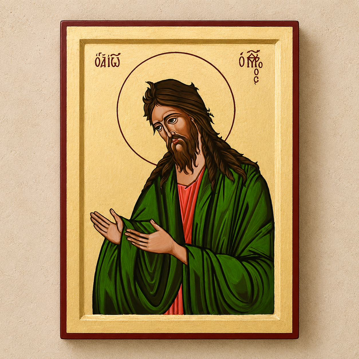 A framed religious icon painting of Saint John on a beige wall. The artwork features a gold background, a red-brown frame, and depicts St John with long brown hair and beard, wearing a green robe over a red tunic