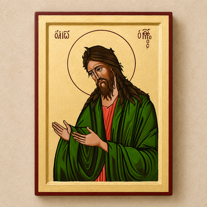 A framed religious icon painting of Saint John on a beige wall. The artwork features a gold background, a red-brown frame, and depicts St John with long brown hair and beard, wearing a green robe over a red tunic