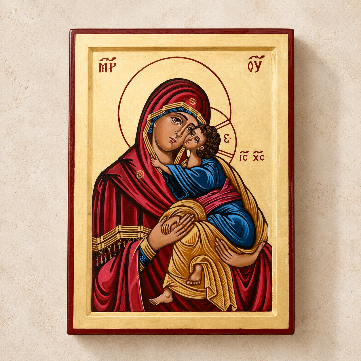 A traditional Eastern Orthodox icon of the Virgin Mary holding the infant Jesus, mounted on a light beige textured wall. The icon features a gold background with a red border, Mary in a deep red robe, and Jesus in blue and gold garments.