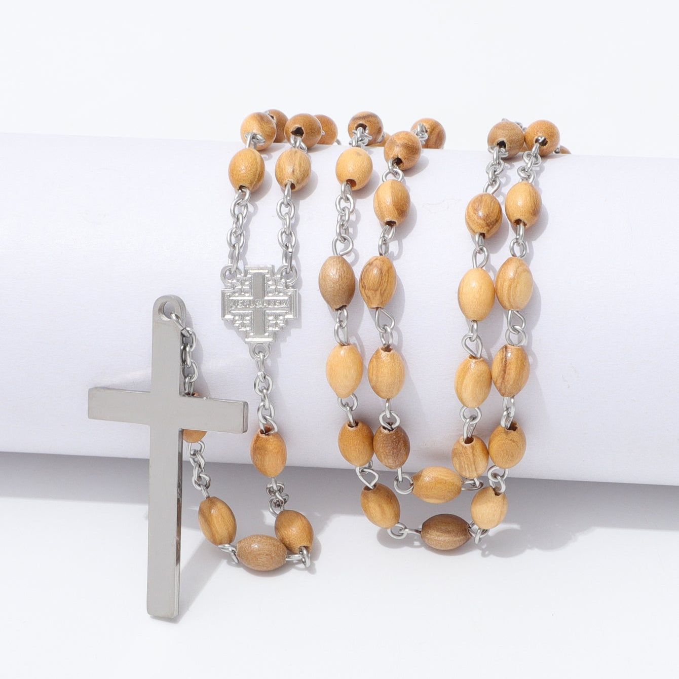 A rosary-style necklace with light tan wooden beads and silver-tone metal links, cross, and centerpiece, displayed on a white cylindrical prop against a white background.