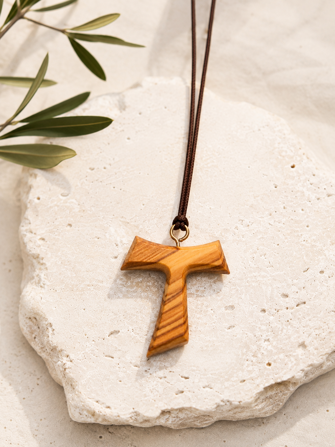 A wooden cross pendant necklace with a natural olive wood grain finish, suspended on a dark brown cord, resting on a textured white stone with green leaves in the corner.