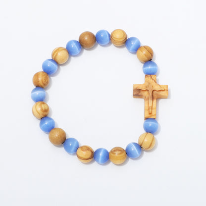 A beaded stretch bracelet featuring alternating light blue and natural wood beads, with a small carved wooden cross on the side, displayed on a clean white background.
