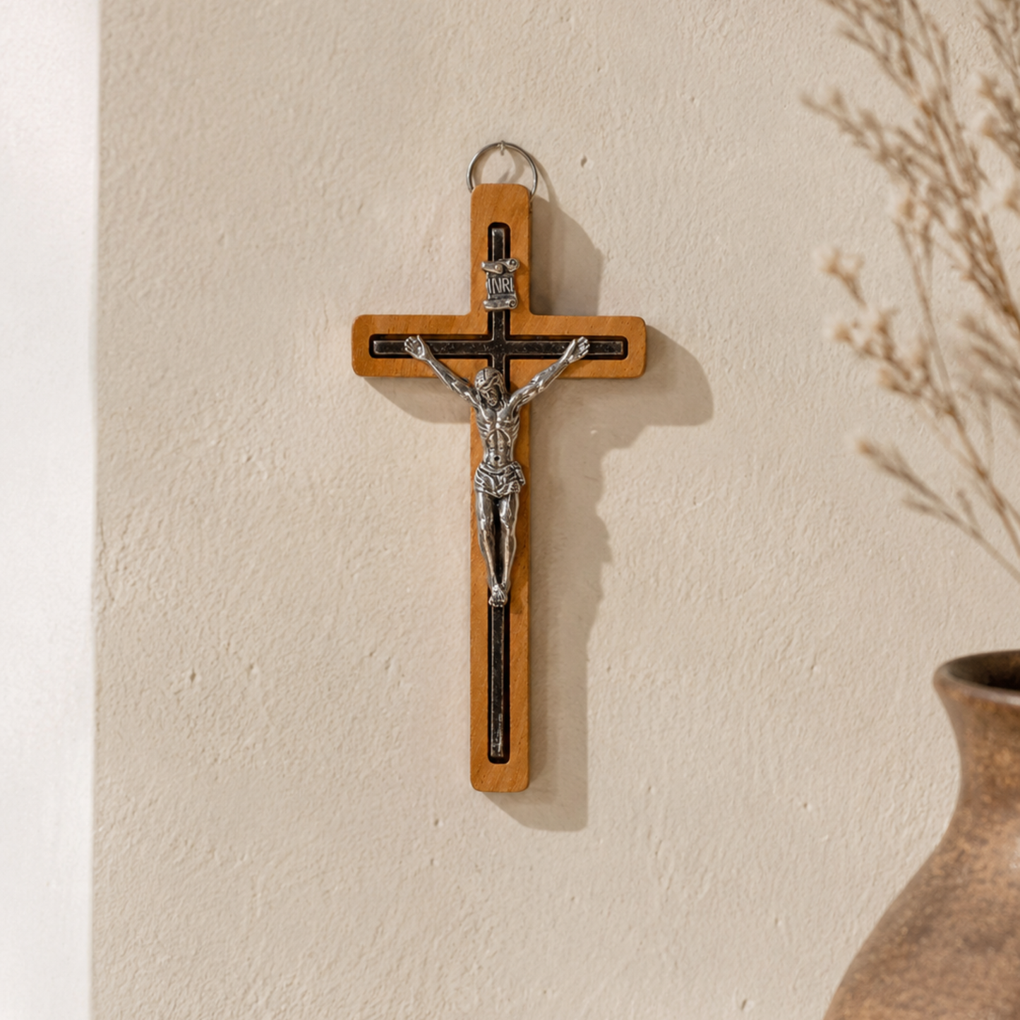 A wooden wall cross with a silver-toned metal corpus of Jesus Christ, hanging on a textured beige wall next to a brown ceramic vase with dried branches.