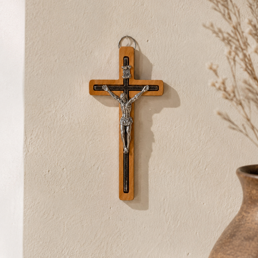 A wooden wall cross with a silver-toned metal corpus of Jesus Christ, hanging on a textured beige wall next to a brown ceramic vase with dried branches.