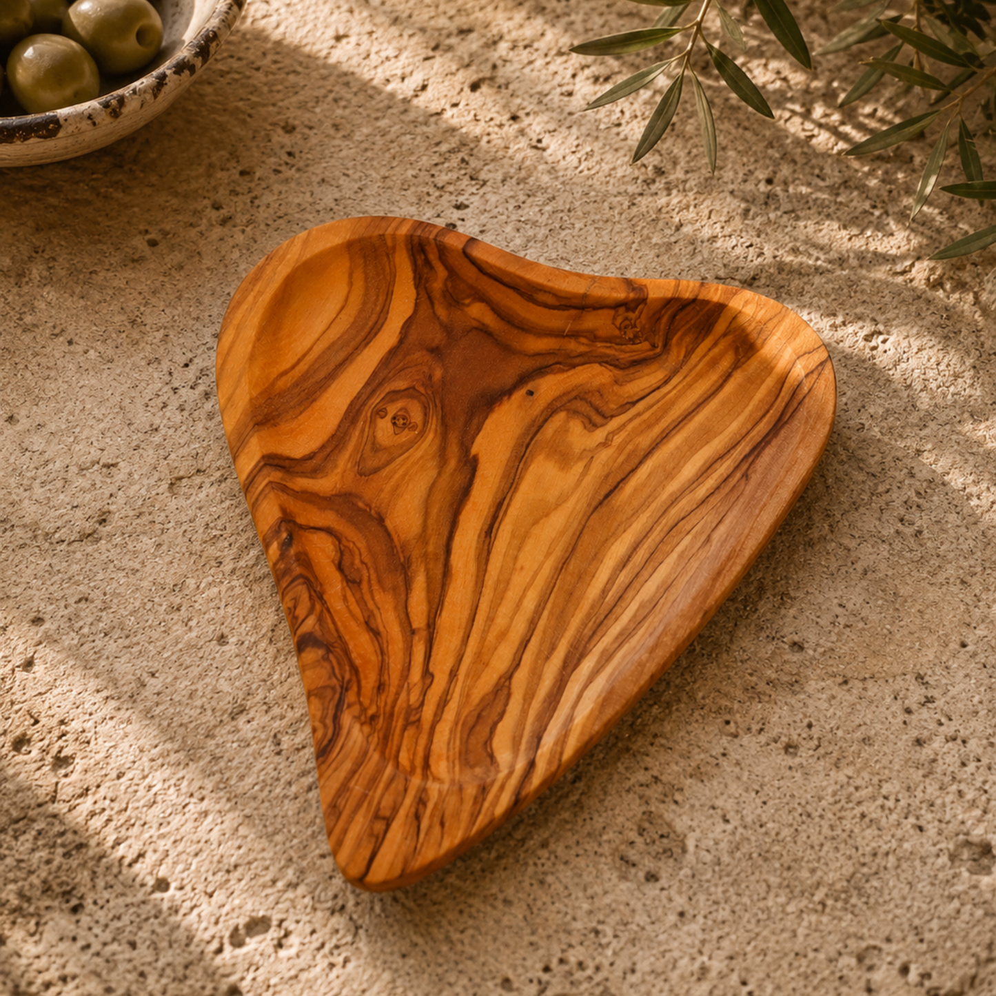 A heart-shaped wooden serving tray with a rich, swirling grain pattern in warm golden-brown and amber tones, resting on a textured sandy surface with soft shadows.