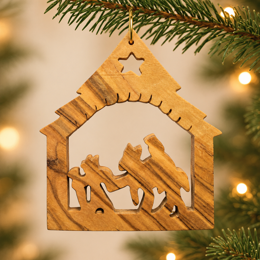 A wooden nativity ornament shaped like a stable, hanging from a pine branch with warm blurred lights in the background.