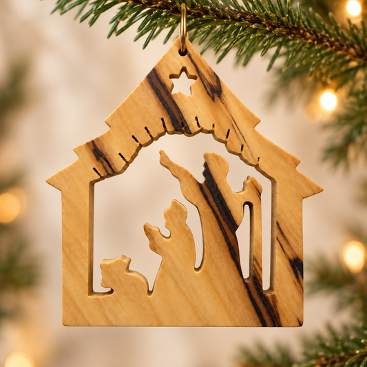 A wooden nativity ornament shaped like a stable, hanging from a pine tree branch with warm bokeh lights in the background.
