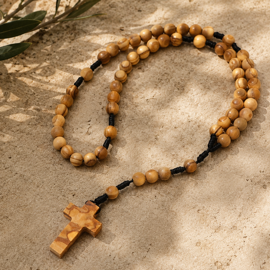 A wooden bead rosary with a matching wooden cross, laid out on a sandy surface with soft leaf shadows.