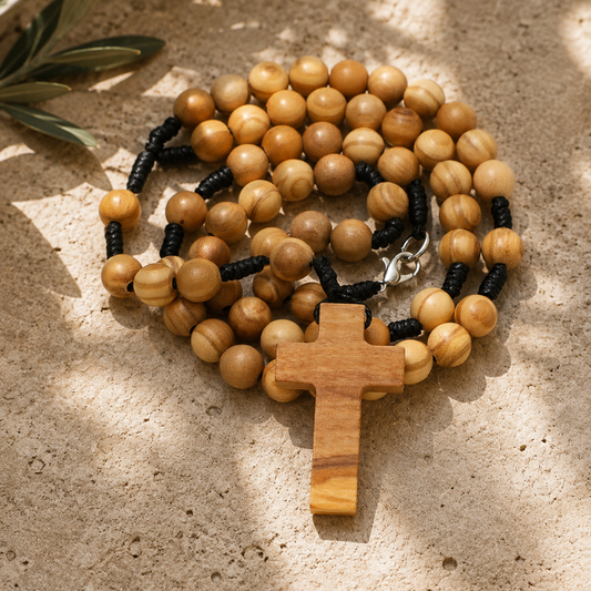 A wooden bead rosary with a wooden cross pendant, arranged on a textured stone surface with dappled sunlight and shadows.