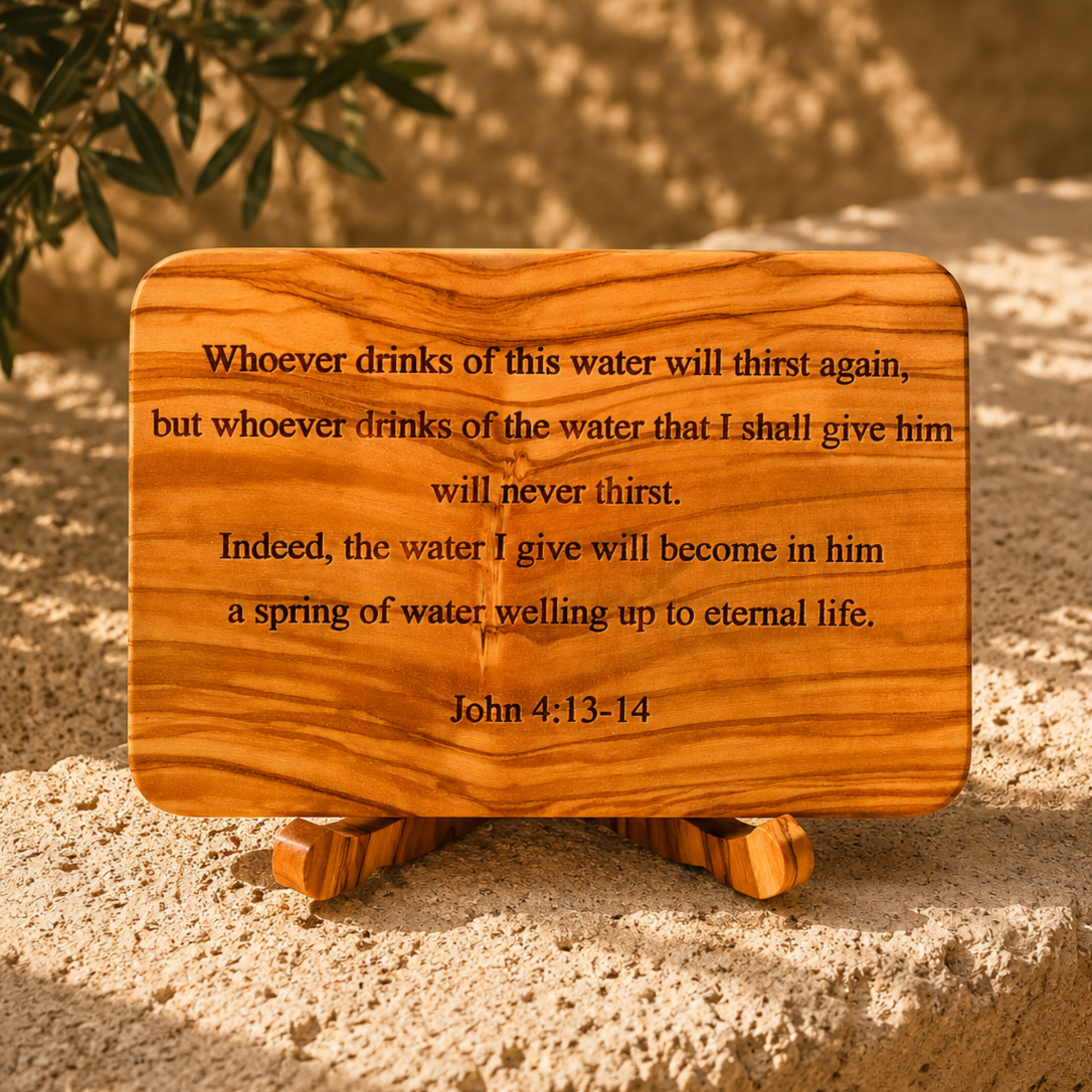A rectangular olive wood plaque with rounded corners and a small matching wooden stand, engraved with the Bible verse John 4:13-14. The plaque is displayed outdoors on sandy ground with soft-focus olive leaves in the background.