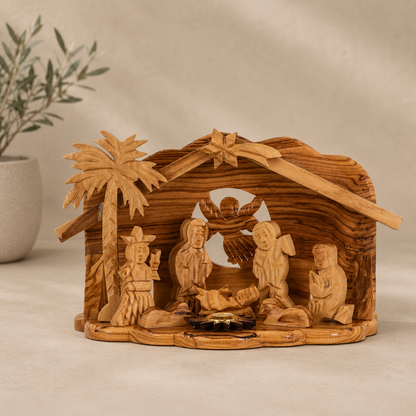 Hand-carved olive wood nativity scene with a stable, palm tree, and multiple figures, displayed on a neutral beige surface with a soft-focus plant in the background.