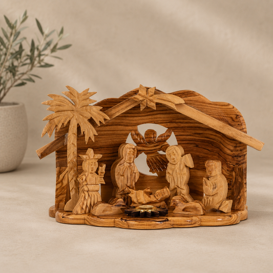 Hand-carved olive wood nativity scene with a stable, palm tree, and multiple figures, displayed on a neutral beige surface with a soft-focus plant in the background.