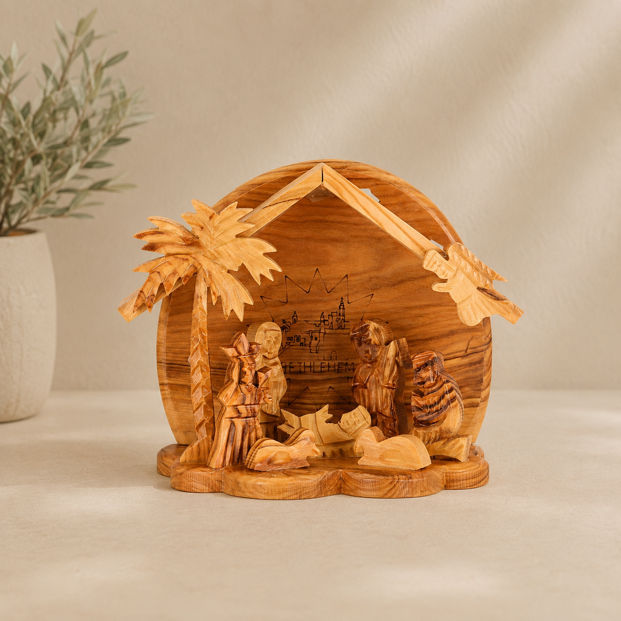 Hand-carved olive wood nativity scene with a thatched-roof stable, palm tree, and small wooden figures, displayed on a light surface with a blurred plant in the background.
