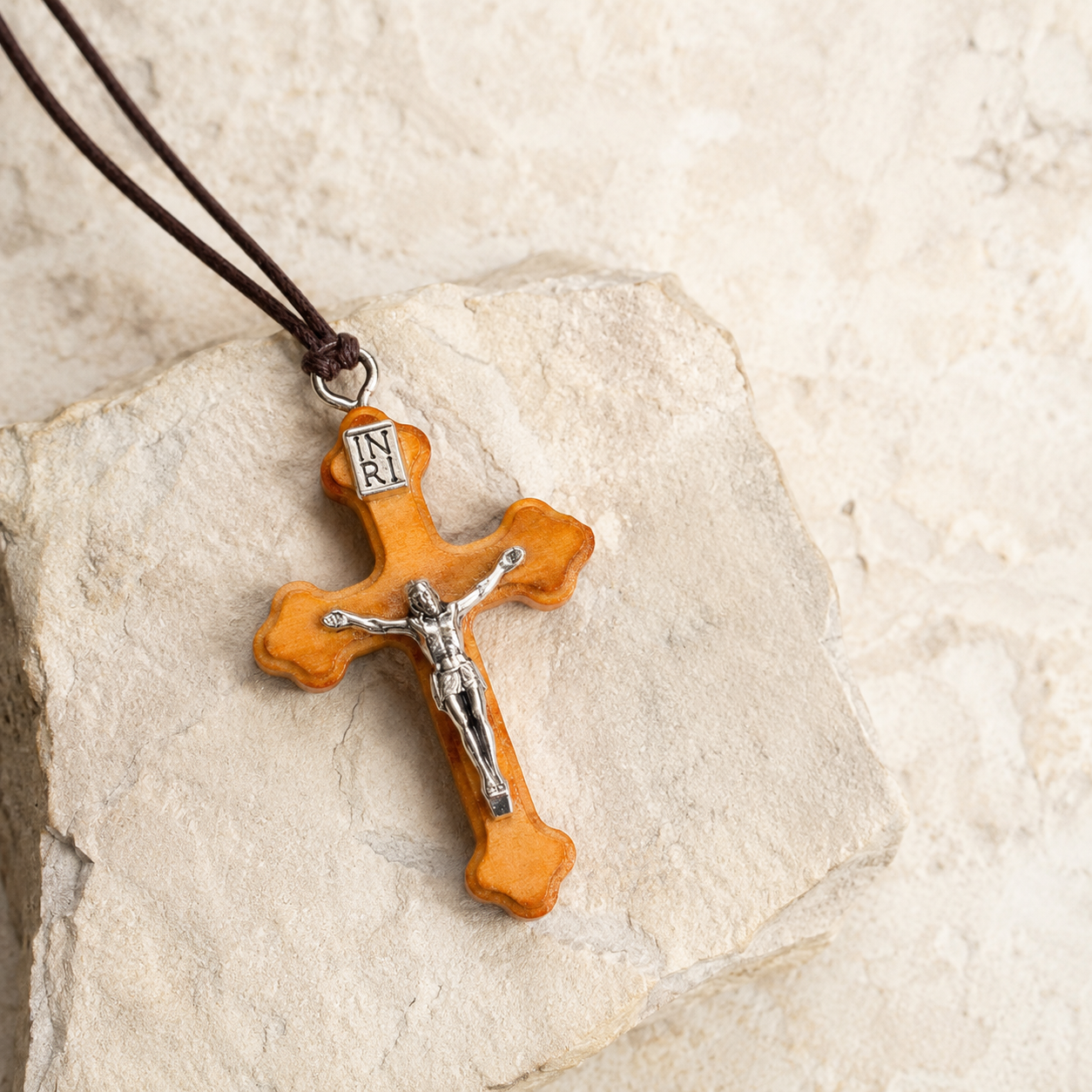A wooden cross pendant necklace with a silver-toned crucifix and a dark brown cord, displayed on a light beige stone surface.
