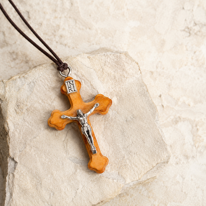 A wooden cross pendant necklace with a silver-toned crucifix and a dark brown cord, displayed on a light beige stone surface.