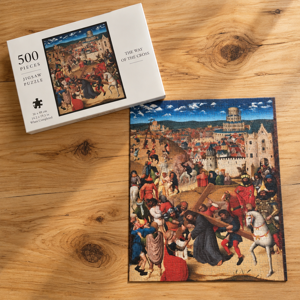 A 500-piece jigsaw puzzle about the Way of the Cross displayed next to its white box on a light brown wooden tabletop. The puzzle is completed and shows a detailed medieval scene with figures carrying the cross, while the box displays the product's front cover art.