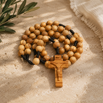 A wooden rosary with light tan beads and a matching wooden crucifix, arranged on a textured beige stone surface with olive leaves and a cream fabric in the background.