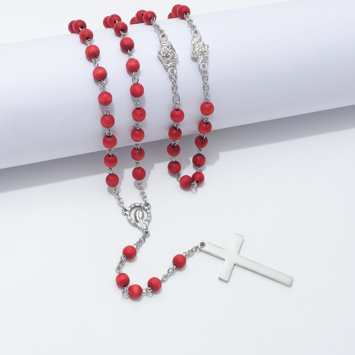 Red beaded rosary with silver-tone metal links, centerpiece, and cross, arranged on a white background with a rolled white cylinder prop.