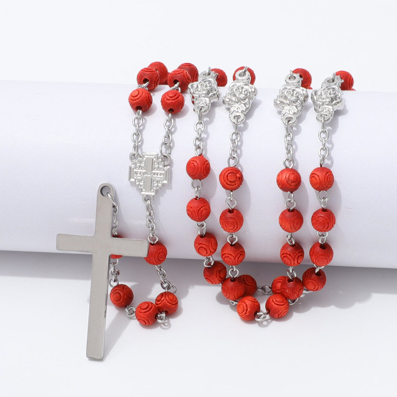 A rosary-style necklace with bright red faceted beads, silver-tone chain links, ornate silver medallions, and a silver cross pendant, displayed on a white cylindrical prop against a white background.
