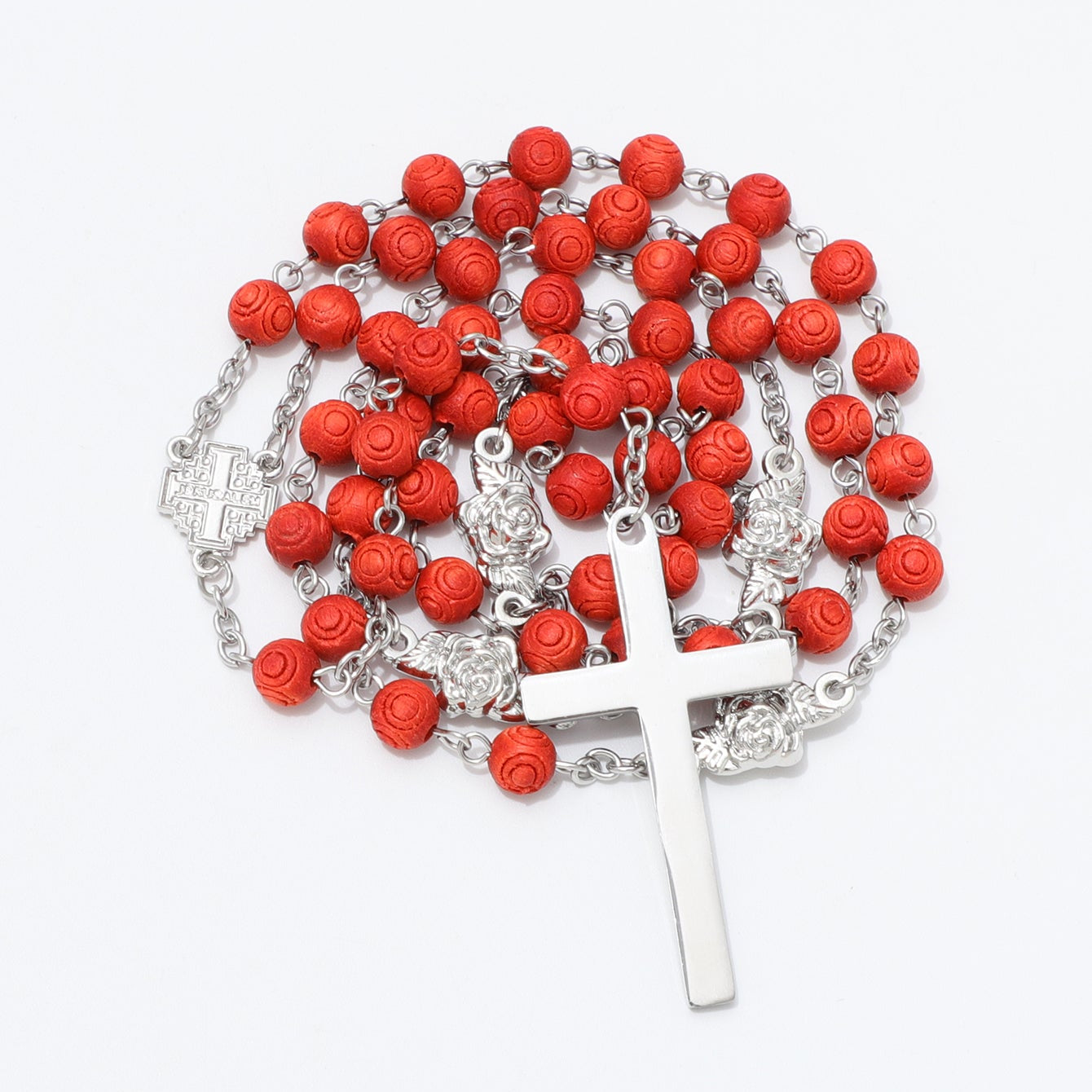 A rosary necklace featuring textured red rose-shaped beads on a silver-tone chain, with a silver-tone crucifix pendant and a small silver-tone medal, displayed on a clean white background.