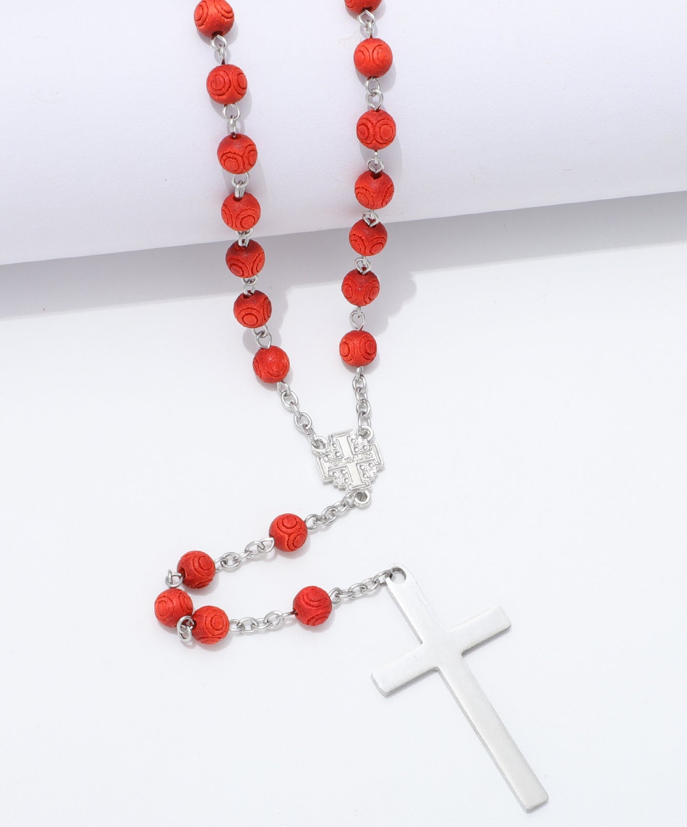 A red and silver rosary necklace featuring textured red beads, a silver chain, a silver centerpiece, and a polished silver cross pendant, displayed on a clean white background.