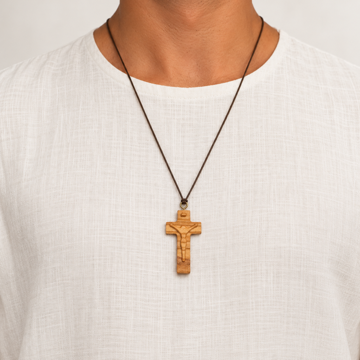 A close-up of a person wearing a wooden crucifix pendant on a dark brown cord necklace, paired with a white linen t-shirt.