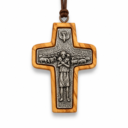 A wooden cross pendant with an olive wood grain finish and a detailed silver-toned metal centerpiece depicting Jesus and sheep, suspended by a brown cord on a white background.
