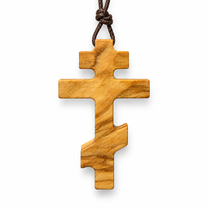 An olive wood Orthodox cross pendant with a natural grain pattern, hanging from a dark brown knotted cord on a white background.
