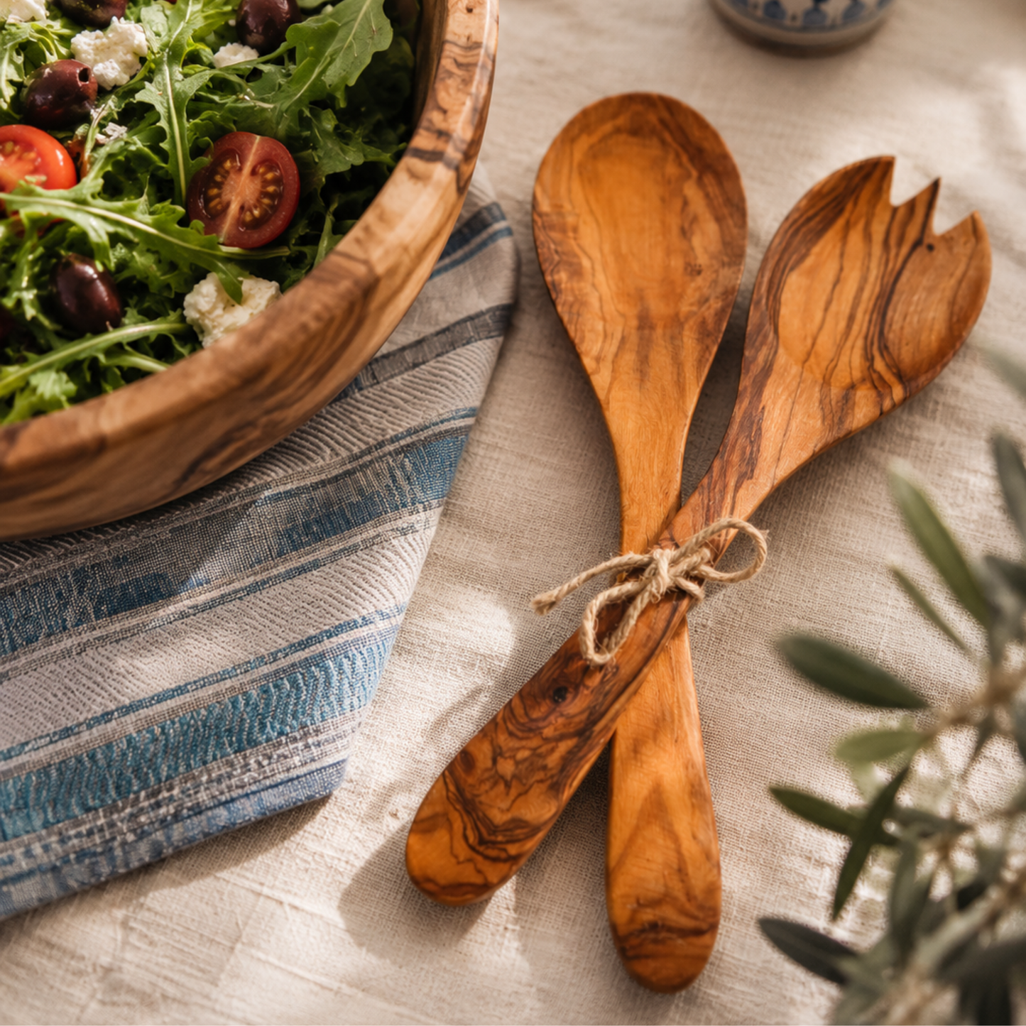Two wooden salad serving spoons tied with twine, placed on a linen tablecloth beside a wooden bowl of salad and a striped cloth.