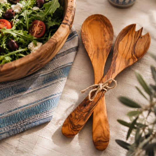 Two wooden salad serving spoons tied with twine, placed on a linen tablecloth beside a wooden bowl of salad and a striped cloth.